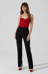 A woman is wearing a red sleeveless top with thin shoulder straps that have small bows on the shoulders. She pairs it with high-waisted black trousers that have a front button and zipper closure. She completes her outfit with red high-heeled shoes.